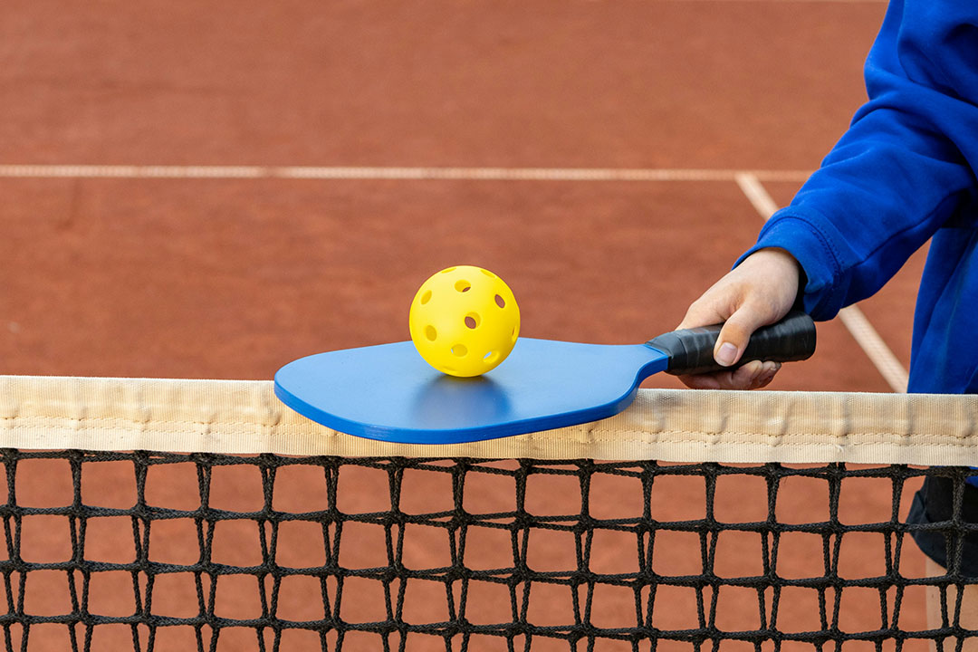 pickleball2_1080x720 Person holding Pickleball bat and ball near tennis net